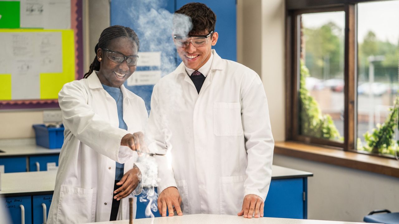 Two students in lab coats conducting a science experiment in classroom.