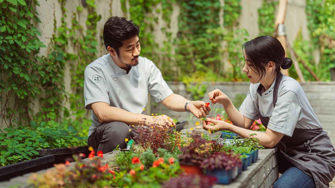 Two chefs working together in a lush outdoor garden setting.