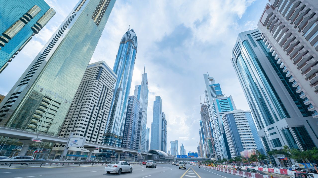 A low angle shot looking up at numerous modern skyscrapers. A wide highway with multiple lanes runs in front of the buildings, with cars driving on it. The cloudy sky suggests daytime. This photo is ideal for urban development projects, travel, and architecture concepts.