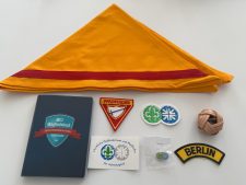 Aufnahme eines Zentrallagers mit Pfadfinder-Accessoires, darunter eine orangefarbene Scout-Halstuch, Abzeichen, Mitgliedsbuch, Kugel, Pins und einen Lederball, die für Pfadfinder-Aktivitäten und Organisationen typisch sind. Die Produkte sind ordentlich auf einem weißen Hintergrund präsentiert und spiegeln die vielfältigen Materialien und Ausrüstungen für Pfadfinderinnen und Pfadfinder wider, die zentral gelagert und verfügbar sind. Perfekt für die Organisation von Pfadfinderaktivitäten und -events.
