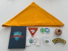 Helles gelbes Halstuch mit Pfadfinderabzeichen und Zubehör auf weißem Hintergrund.