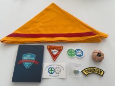 Großes gelbes Pfadfinderhalstuch mit rotem Streifen und einzeln liegende Abzeichen, Sticker, Bälle und ein Mitgliedsbuch, die sich auf Pfadfinder- und Zentrallager-Artikel für die Jugendarbeit beziehen.