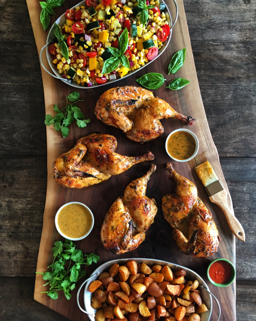 Two grilled piri-piri chickens cut in half and on a large wooden board. Served with roasted potatoes and a corn and tomato salad.