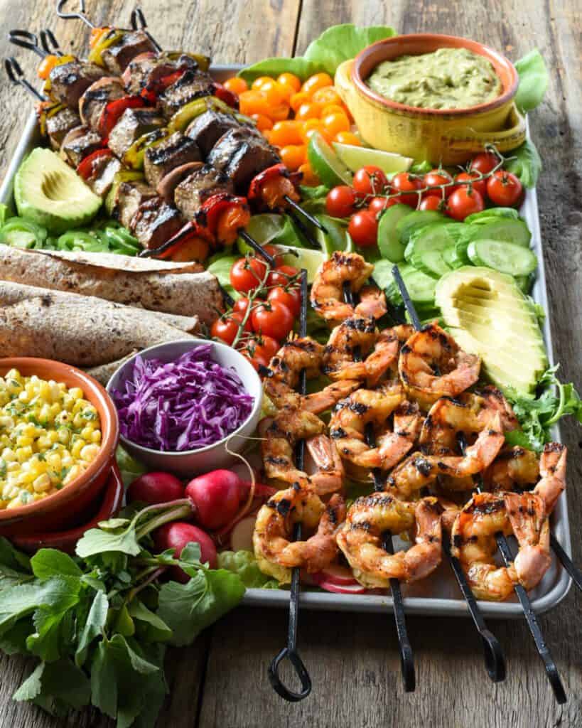 A large and colourful surf and turf shrimp and beef skewers on a platter, alongside grilled tortillas, spicy guacamole, tomatoes, avocados, sliced jalapenos, grilled corn and tomato salad, lime wedges and fresh chopped cilantro.