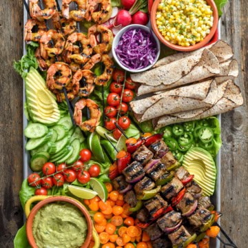 A surf and turf platter of beef and vegetable skewers, grilled shrimp, tortillas and a large selection of colourful toppings.
