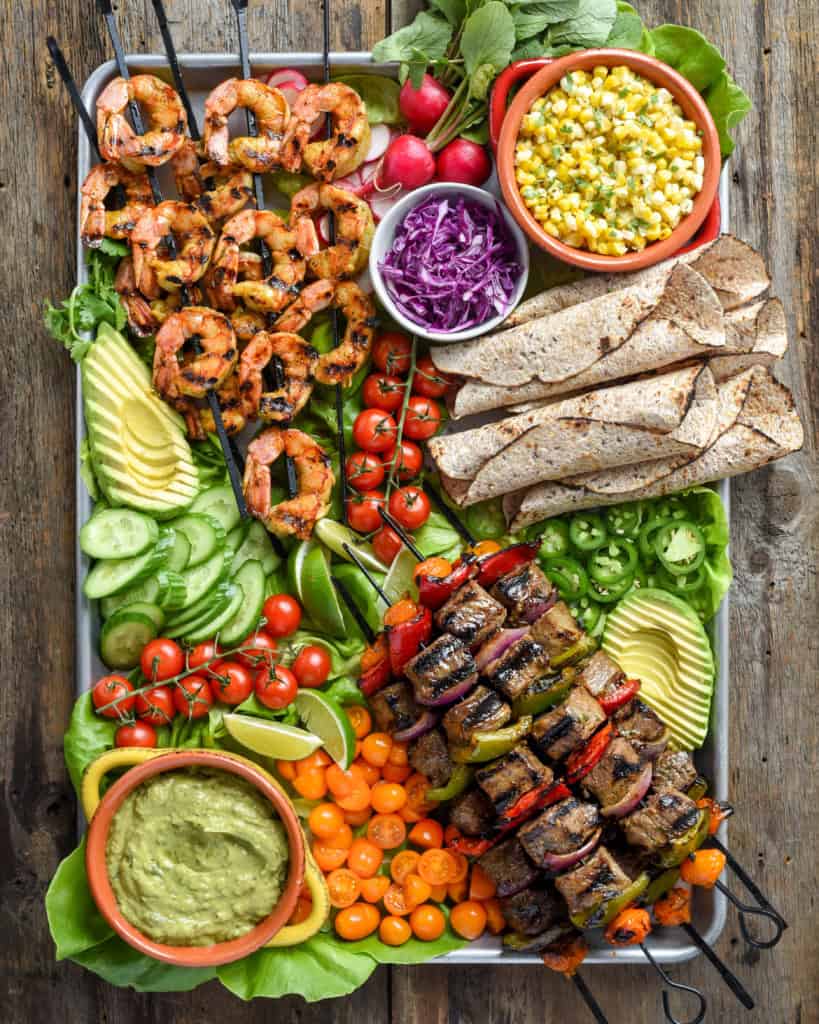 A surf and turf platter of beef and vegetable skewers, grilled shrimp, tortillas and a large selection of colourful toppings.