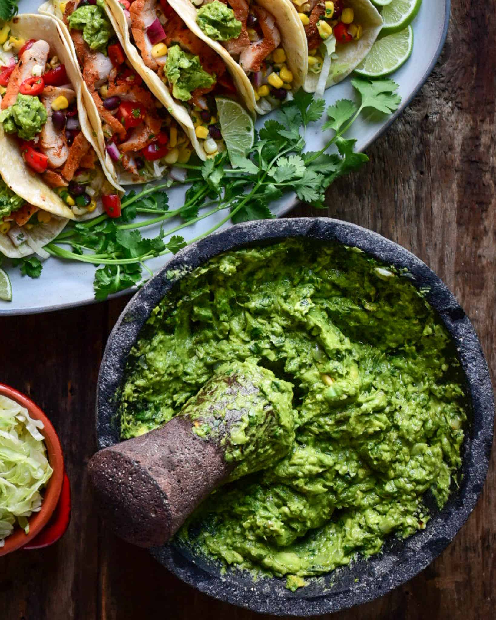 A large molcajete with freshly made grilled guacamole with a plate of tacos in the background.