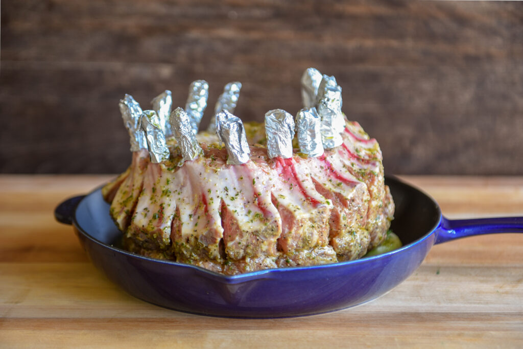 Raw crown roast of pork in a blue cast-iron pan, marinated with tin foil on the bones.