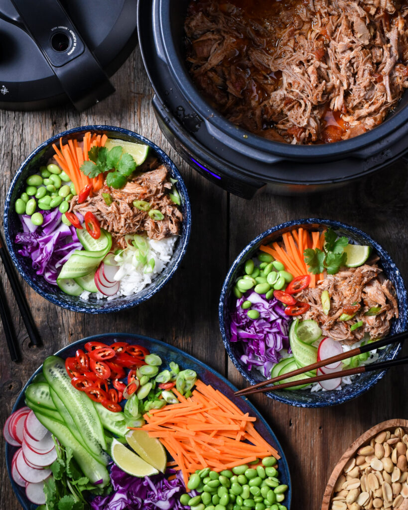 Top down view of a table with a crock-pot full of a shredded pork shoulder. Two blue bowls filled with rice, pork, carrots, radishes, cucumber, cabbage, red peppers and beans.