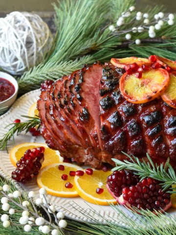 Side view shot of a glazed ham sliced garnished with pomegranates, rosemary and sliced oranges. Surrounded by Christmas decorations and a bowl of cranberry sauce.