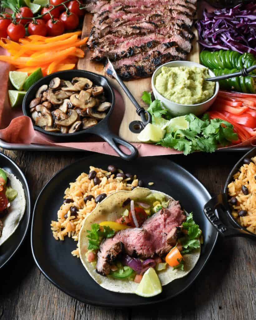 A medium rare slice of flank steak in a taco with bright toppings infront of a large taco platter.