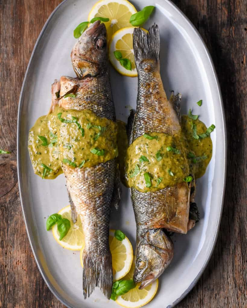 Two grilled branzinos on a plate with lemon slices and jalapeño-basil vinaigrette.