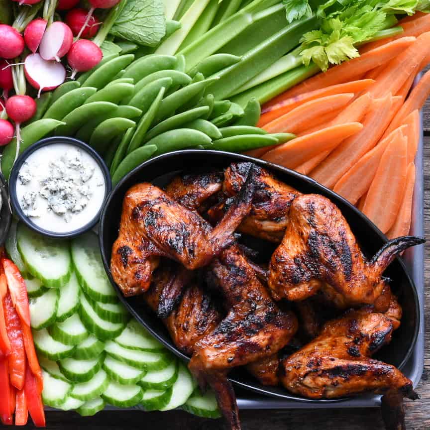 An oval bowl of grilled whole chicken wings on a platter with a veggies including celery, snap peas, radishes, cucumber, carros and two bowls of dip.