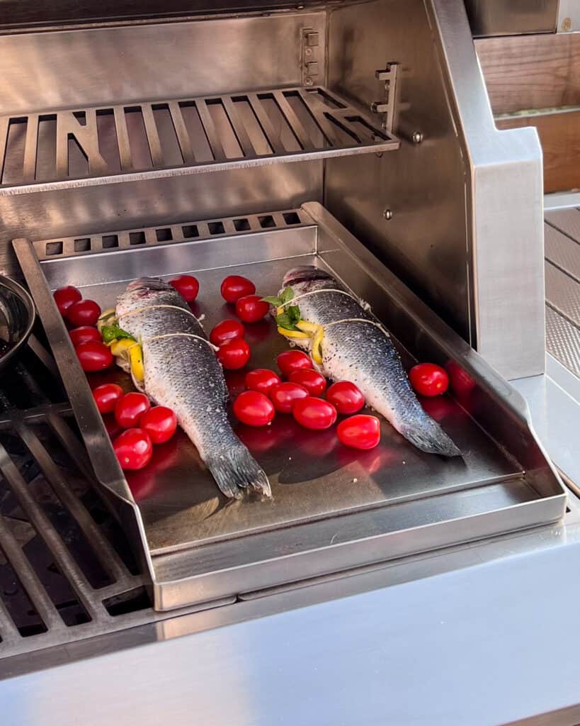 Two whole branzino fish on a grill griddle being cooked with cherry tomatoes.