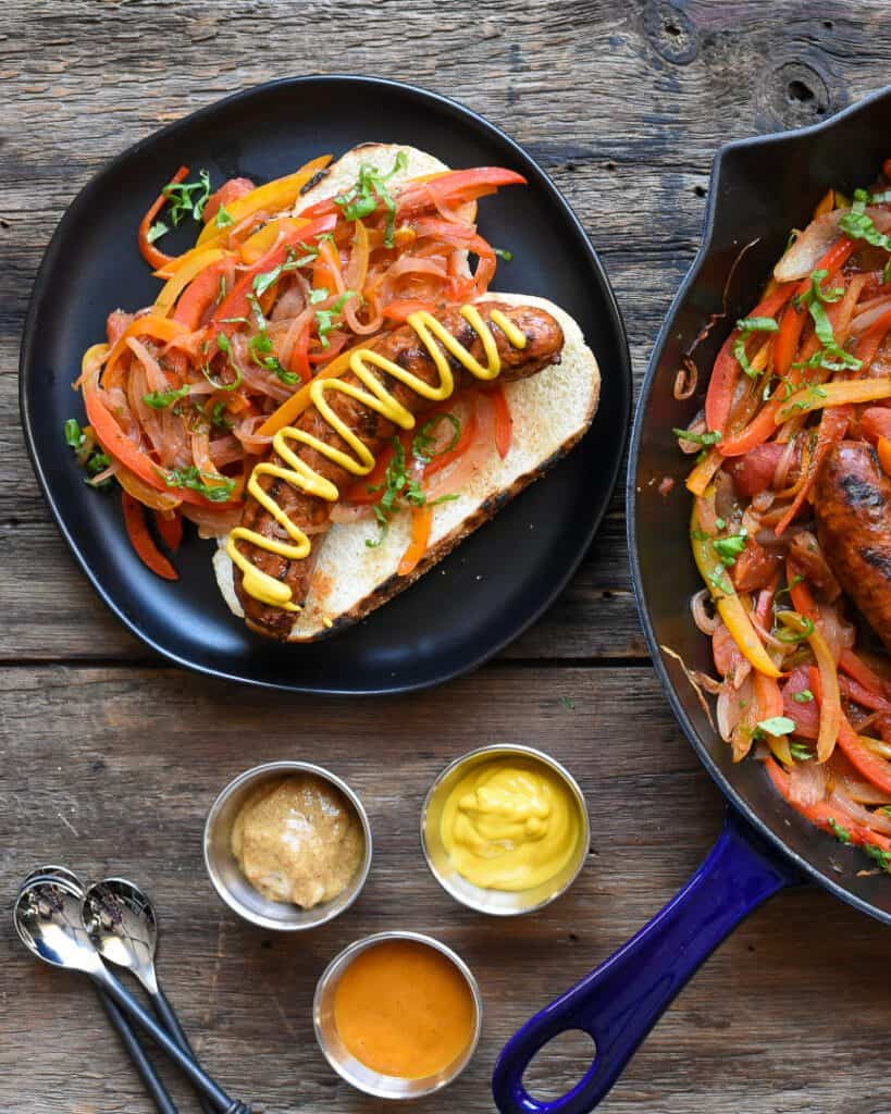 Ontario pork hot Italian sausages with peppers and onions in a blue cast iron skillet and on a bun on a black plate. Three bowls of mustard.