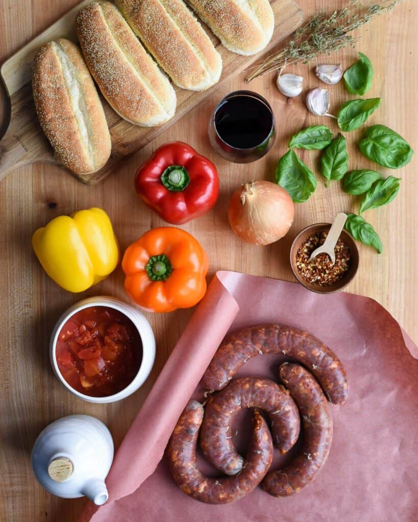 Ingredients laid out including hot Italian sausages, peppers, basil, sausage buns, garlic oil, chili flakes, wine, onion, tomato sauce and olive oil.