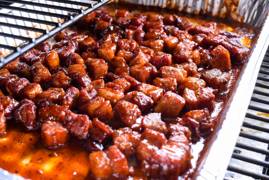 Pork cubes in aluminum pan with sauce in a smoker grill.