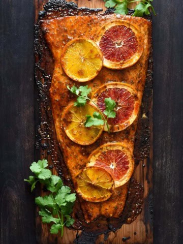 A cooked salmon fillet on a cedar plank with orange slices and cilantro.