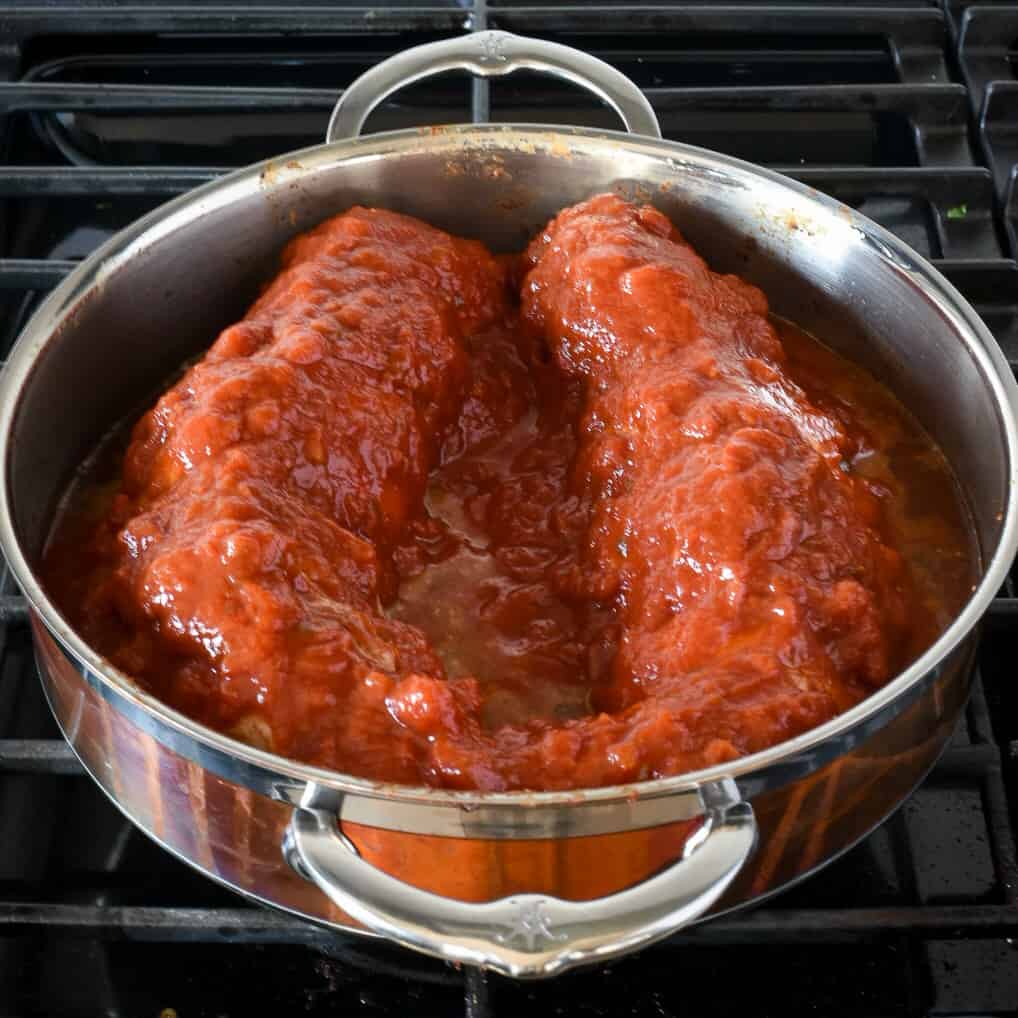 Two pork tenderloins simmering in a tomato sauce in copper pot with two handles.
