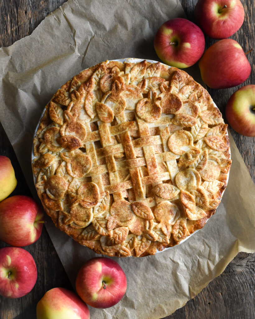 A baked apple pie, decorated with lattice and cut out leaves crust. The pie is surrounded with several Ambrosia Apples.