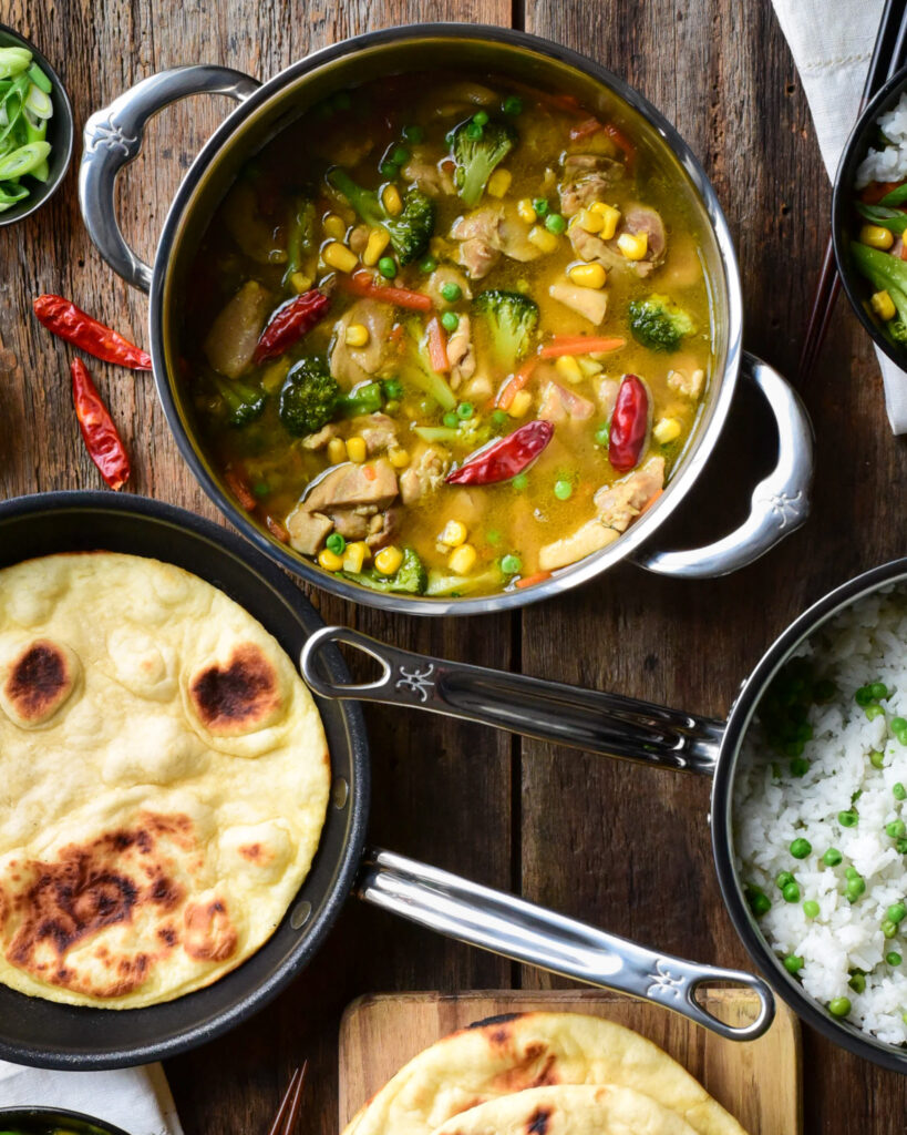 A pot of Thai Green curry chicken, a pot of rice and peas, skillet flatbread and two bowls filled with the curry, rice and garnishes.