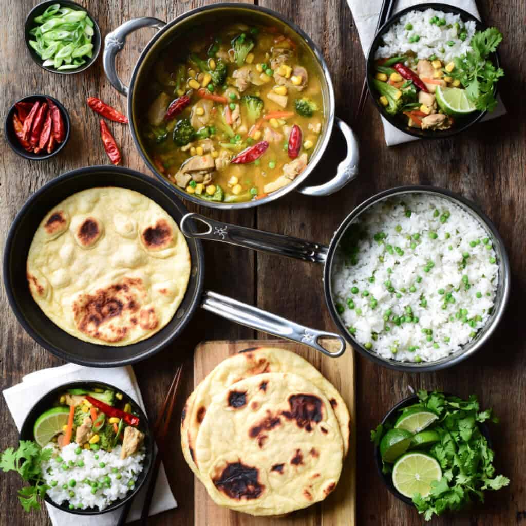 A pot of Thai Green curry chicken, a pot of rice and peas, skillet flatbread and two bowls filled with the curry, rice and garnishes.