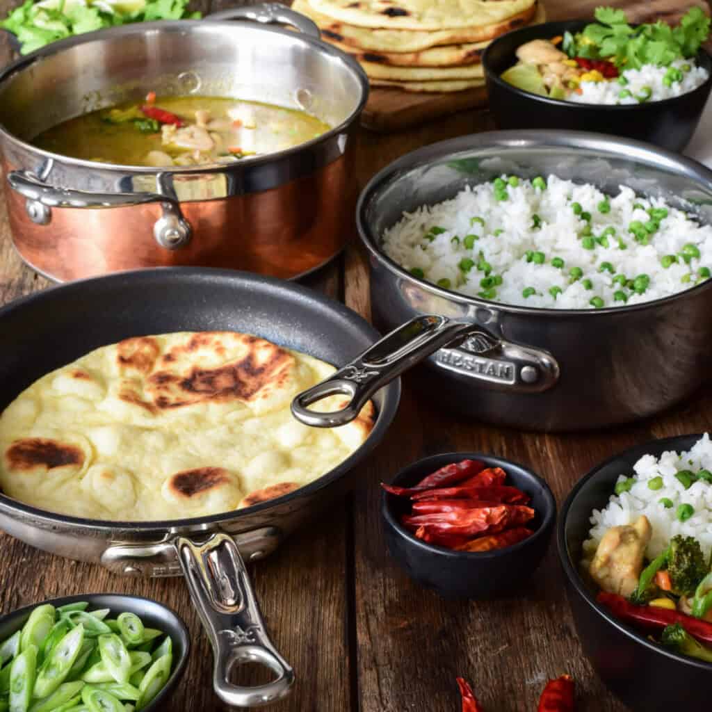 A copper pot filled with green curry, a pot of rice and peas, and a skillet with flatbread.