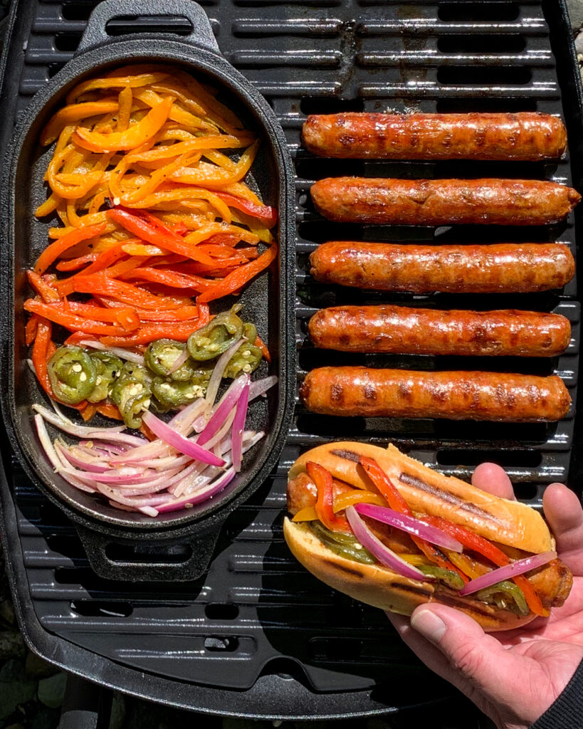 A bbq with grilled spicy Italian sausages, a cast iron pan with vegetables and someone holding a sausage on a bun fully loaded.