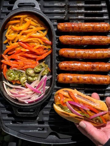 A bbq with grilled spicy Italian sausages, a cast iron pan with vegetables and someone holding a sausage on a bun fully loaded.