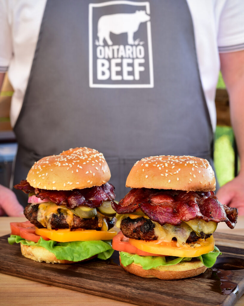 Zimmy wearing an Ontario Beef apron with 2 signature burgers on a wooden board.