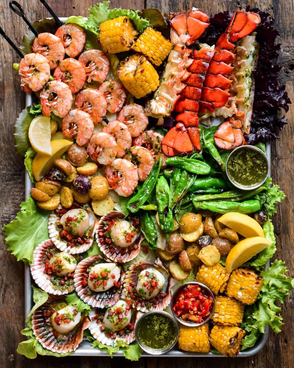 A large platter filled with grilled skewered shrimp, scallops in the shell, lobster tails, grilled corn on the cob and other fresh veggies.
