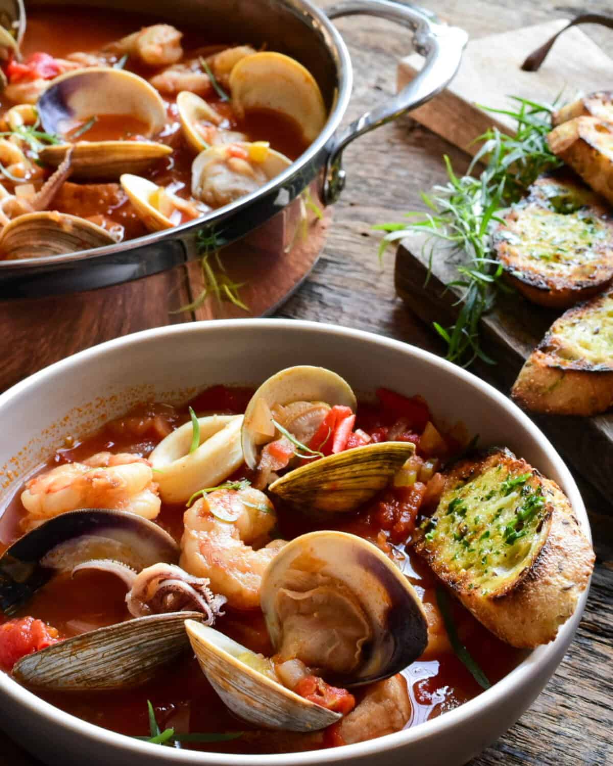A close up of a bowl of cioppino with more stew in a copper pan and served with garlic-herb toast.