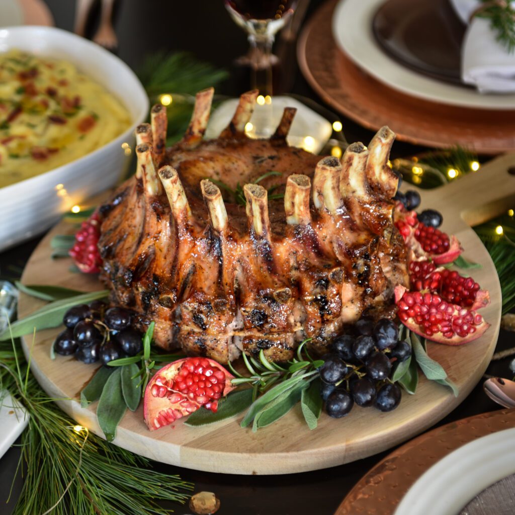 Plated crown roast of pork garneshes with herbs, grapes, and pomegranites on the dinner table. Table is set with dishes and wine glasses.