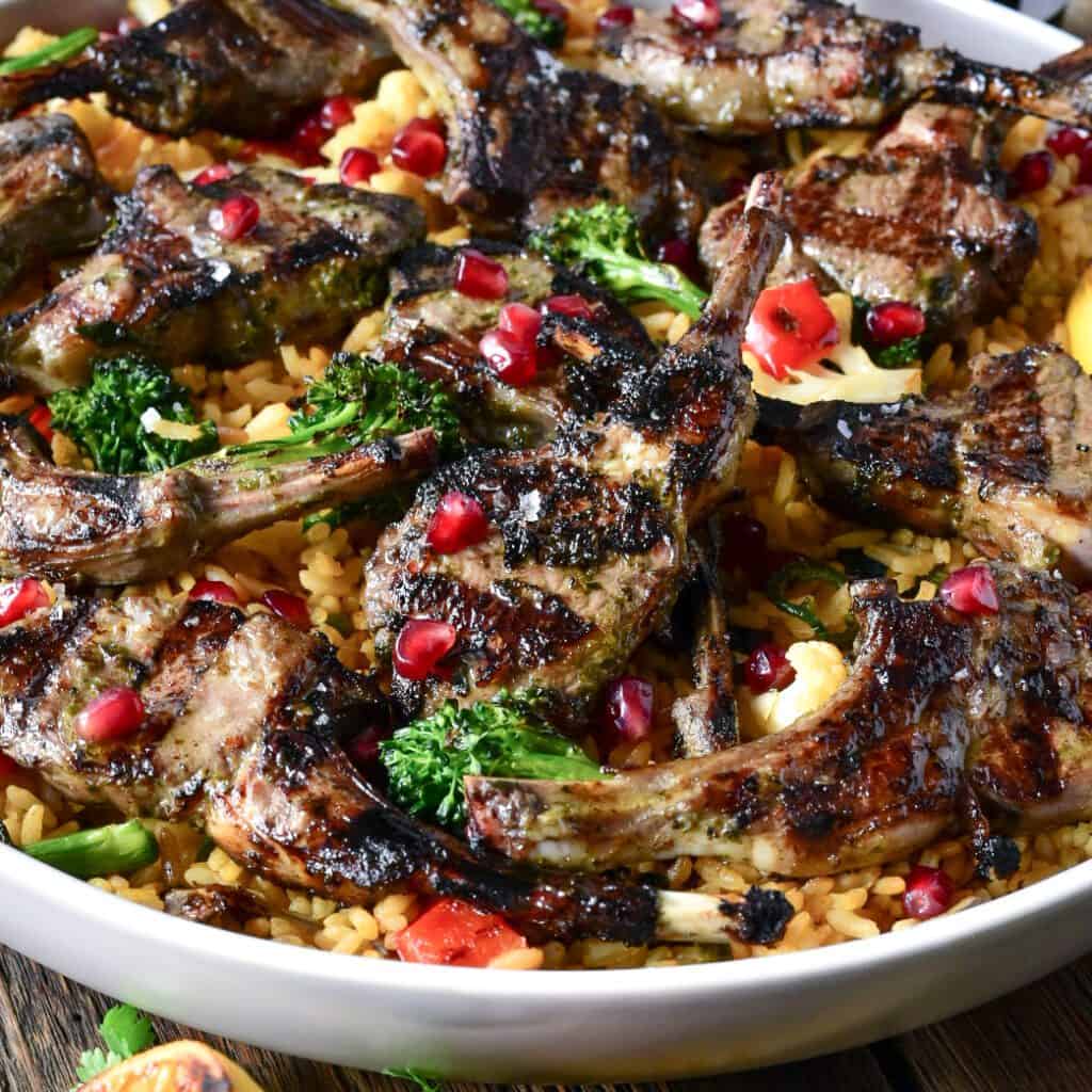 A close up image of a round platter of grilled lamb lollipops served over mixed vegetable rice and pomegranate arils.