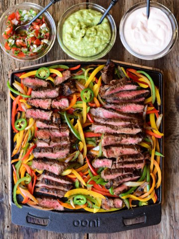 Slices of steak are laid on top of cooked peppers and onions on the Ooni grizzler plate. Served with three sauces.