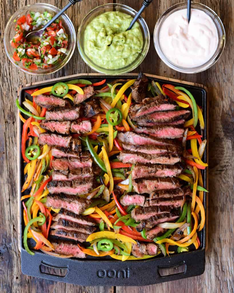 Slices of steak are laid on top of cooked peppers and onions on the Ooni grizzler plate. Served with three sauces.