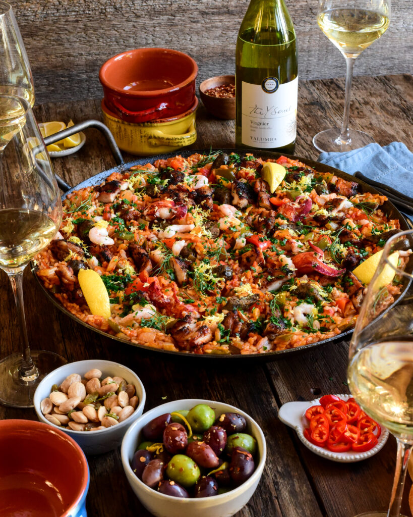 A large, colourful pan of paella filled with seafood, poultry, rice and veggies. This pan is surrounded by a bottle of Viognier, glasses of wine, nuts, olives, lemons and bowls.