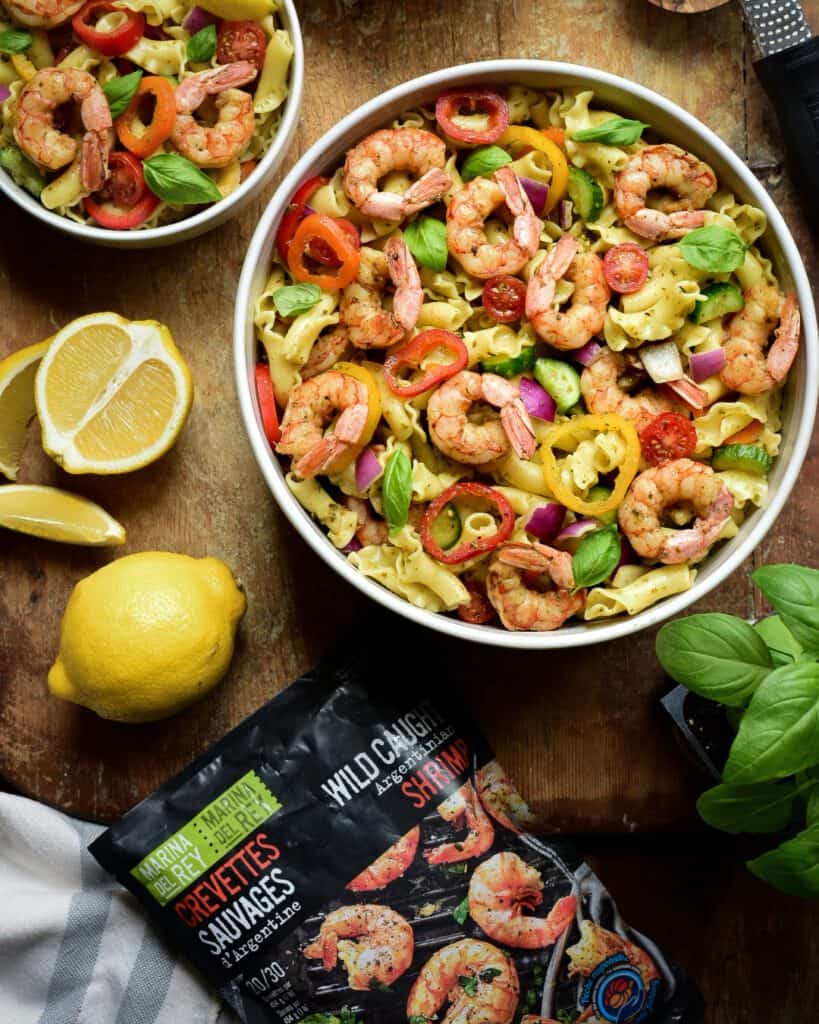 A top down image of a large and two smaller bowls of a Wild Argentinian Shrimp Pesto Pasta Salad on a wooden board. Basil, lemon's and Parmesan cheese.
