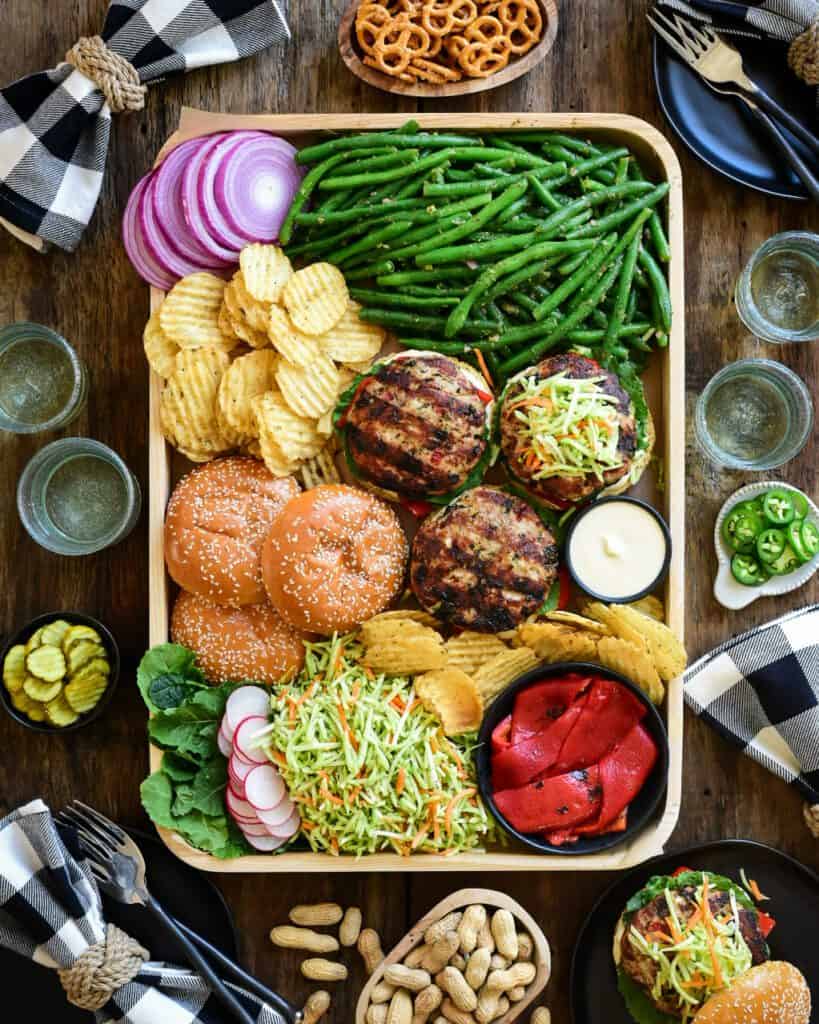A tray of open-faced turkey kale & feta burgers, buns green beans, sliced red onions, chips, dip, broccoli slaw and roasted red peppers. Dishes, glasses, plates and glasses surround this tray.