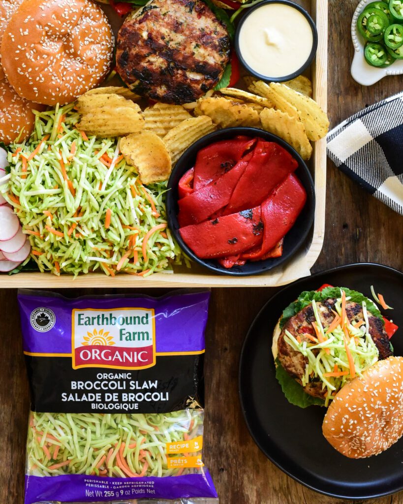 A top down image of a tray filled with burgers, roasted red peppers chips, sauce and Earthbound Organic Broccoli Slaw with Cider Dressing.