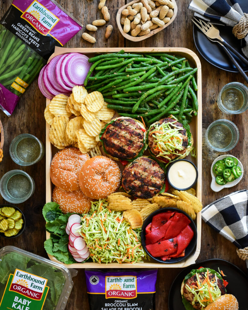 A tray of open-faced turkey burgers, buns green beans, sliced red onions, chips, dip, broccoli slaw and roasted red peppers. Packages of baby kale, broccoli slaw and green beans surround this tray.