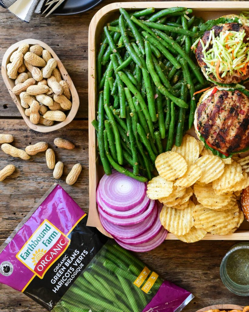 A top down image of a tray filled with Earthbound Green Beans with Lemon, Dill, Dijon Dressing with turkey burgers, ripple chips, sliced red onions. Outside of the tray is the package of Green Beans and a bowl of peanuts. 