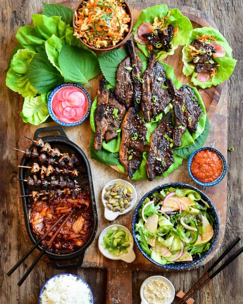 A Korean-Style Barbecue Beef Short Rib Lettuce Wraps spread on a board with coleslaw, salad, grilled mushrooms and kimchi.