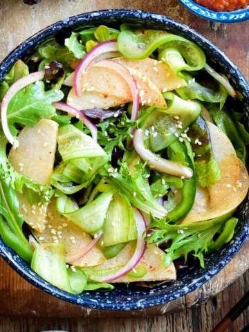 A top down image of a Korean Green Salad with ribbons of cucumbers, slices of Asian pears, onions lettuce, onions and sprinkled with sesame seeds.