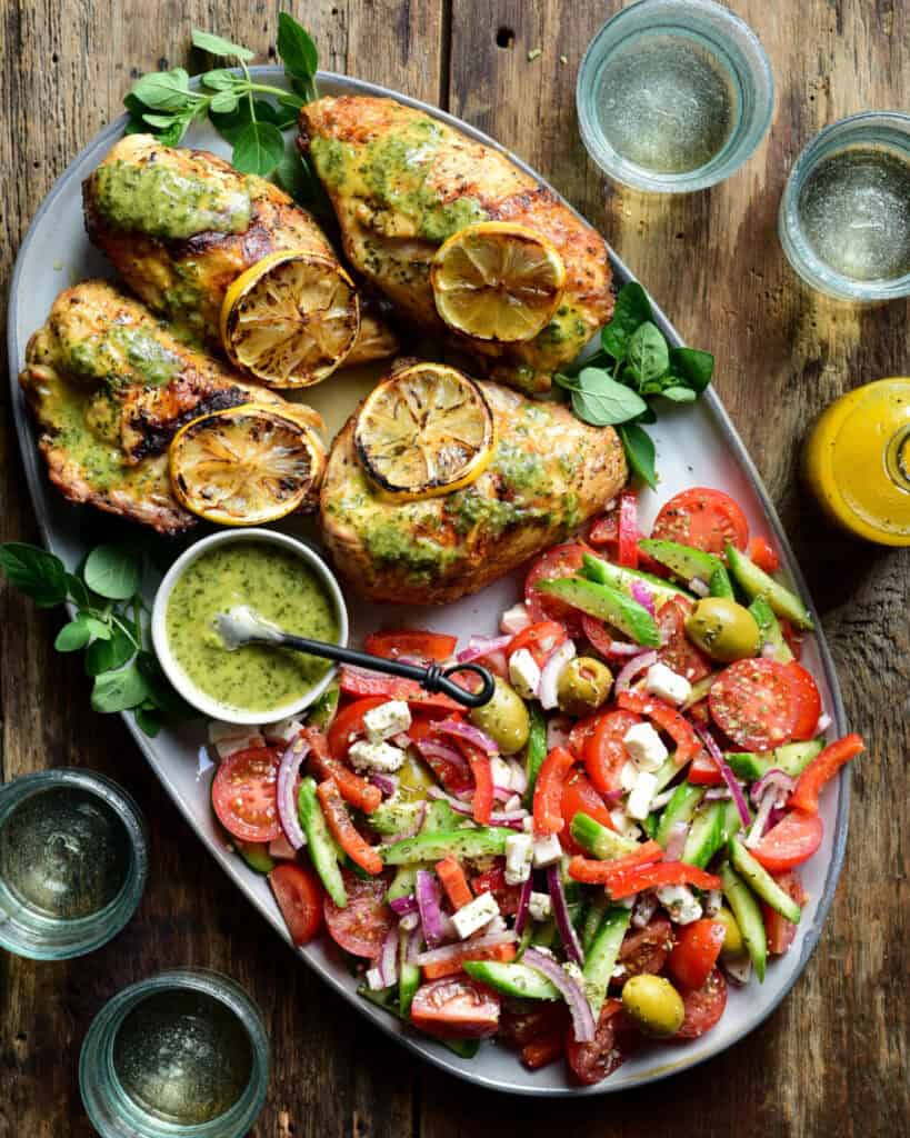 An oval platter with four grilled chicken breasts with grilled lemon slices and fresh oregano on one side and a village salad on the other side. A bowl of green sauce is also in this platter.