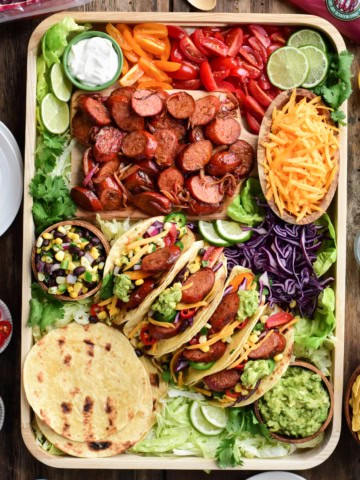 A top down image of a very colourful tray of Bavarian Smokie tacos. Along with the four made up tacos there is a bowl of shredded cheese, a bowl of corn & black bean salsa, a bowl of guacamole, tortillas, sliced grilled sausages, limes, chopped tomatoes and shredded lettuce.