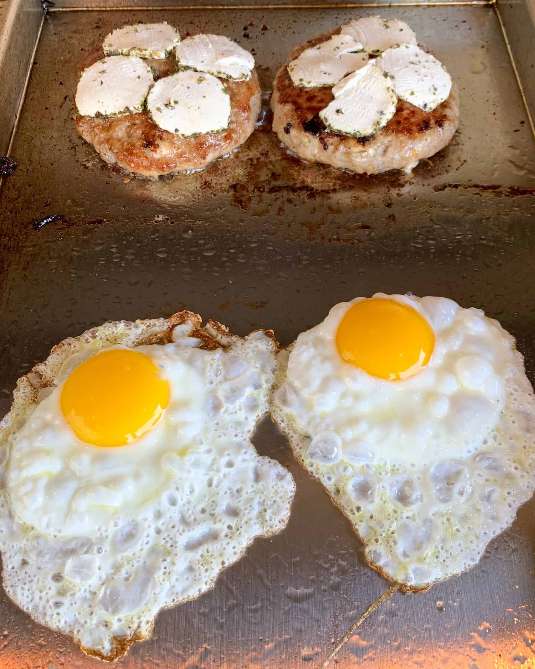 Two turkey burgers topped with goat cheese and two bubbling fried eggs on the griddle.