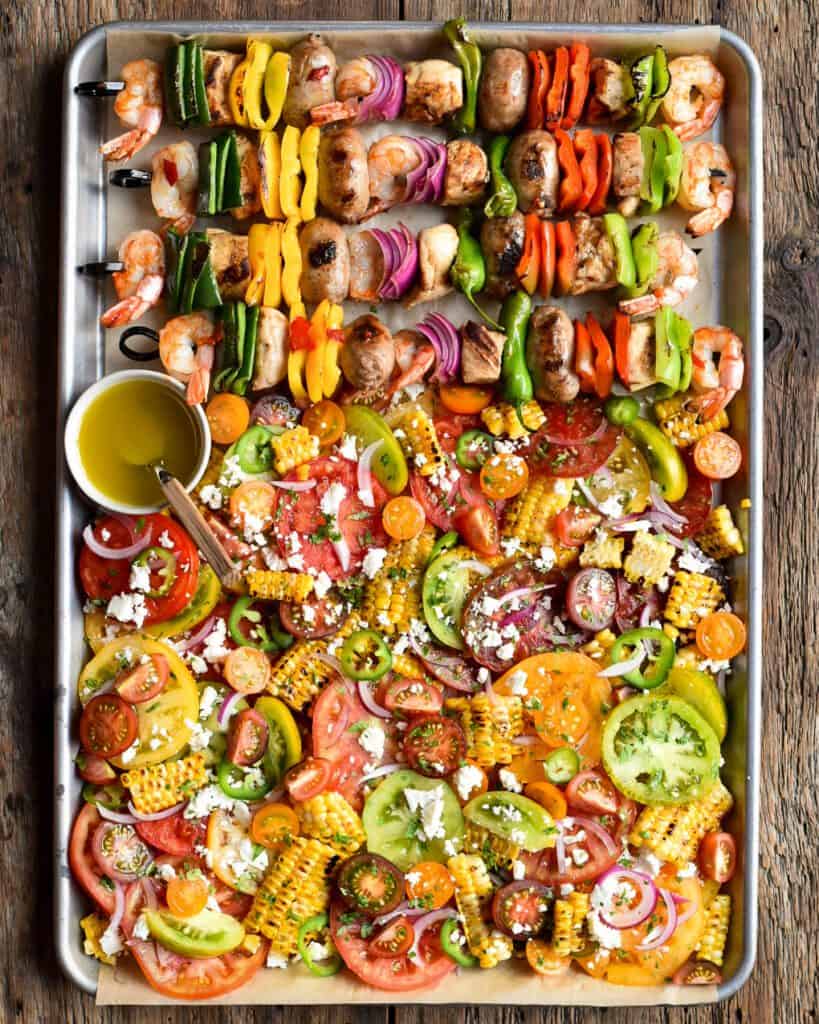 A tray half filled with shrimp, sausage and veggie skewers, and a tomato and grilled corn summer salad.