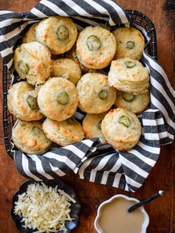 An image of a basket of jalapenos biscuits nestled in a black and white striped tea towel. There is a dish of gravy and a plate of shredded cheese next to this basket.