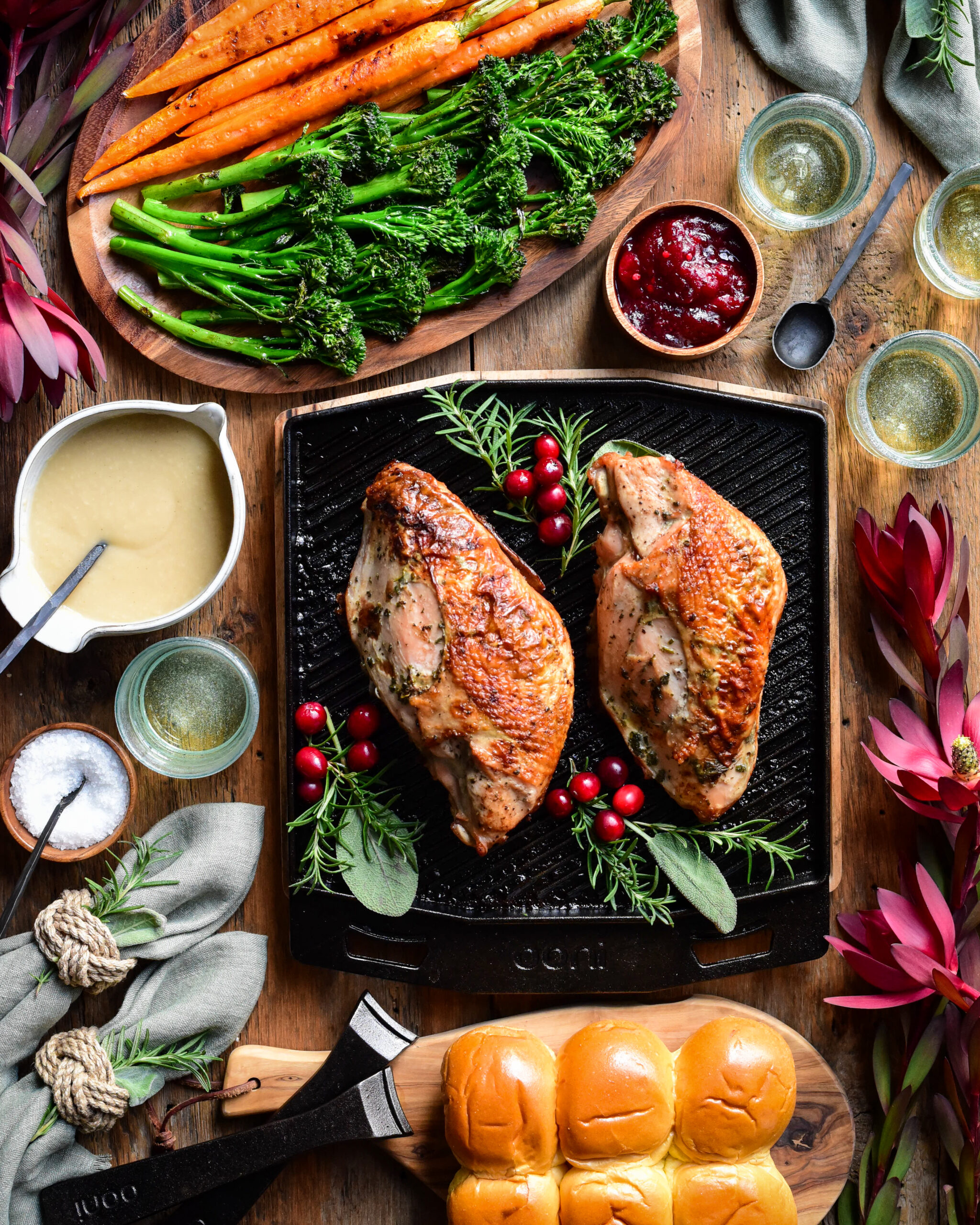 A top down image of two cooked turkey breasts on a skillet with herbs and cranberries. Broccolini, carrots, gravy, buns, cranberry sauce, wine glasses and florals surround this skillet.